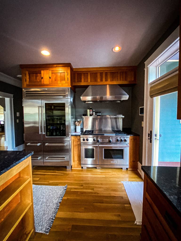 A modern kitchen with wooden cabinets and stainless steel appliances featuring a refrigerator, double oven, and stove. A vent hood is mounted above the stove. The countertops are dark, and the floor is light hardwood. Two rugs are placed on the floor.