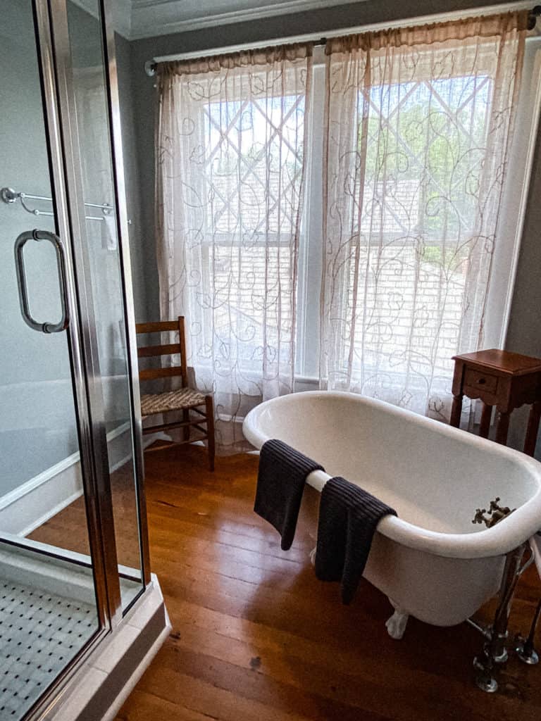 A bathroom with a clawfoot bathtub draped with two dark towels, a glass-enclosed shower, wooden flooring, a wooden chair, a small wooden side table, and two large windows covered with sheer, patterned curtains.