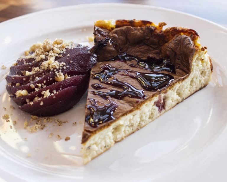 A dessert plate featuring a slice of cheesecake topped with chocolate sauce and a side of poached pear garnished with crumbled nuts. The cheesecake has a browned top and a rich, creamy texture. The pear is sliced and glazed, complementing the sweet treat.