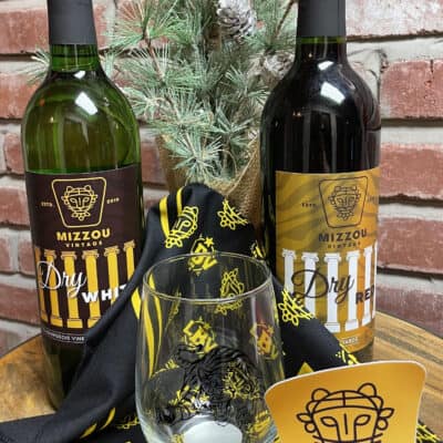 Gift Set: M I Z Dry includes two bottles of Mizzou wine (Dry White and Dry Red), accompanied by a Mizzou-themed glass, napkin, and a decorative pine centerpiece. It is beautifully arranged with a Mizzou card featuring a lion's face logo in front of a brick wall background.