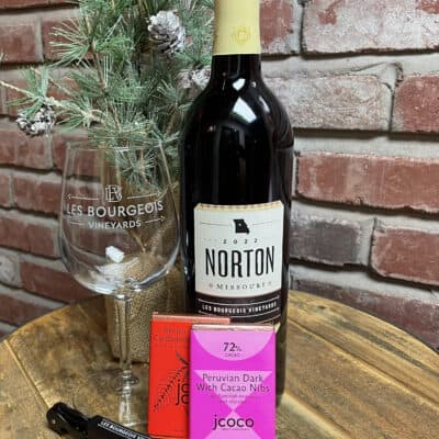 The "Gift Set: Norton & Chocolate" from Les Bourgeois Vineyards includes a bottle of 2021 Norton red wine, a branded wine glass, and a small evergreen decoration, accompanied by two jcoco chocolate bars labeled "Peruvian Dark with Cacao Nibs," along with a wine opener.