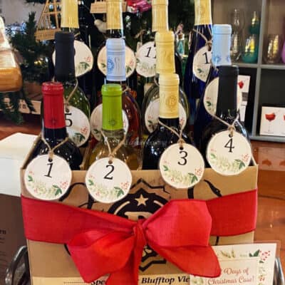 A festive cardboard box, available under the product name "12 Days of Christmas Case Sales," contains 12 bottles of wine, each adorned with circular tags numbered 1 through 12. The box is decorated with a large red bow, and holiday-themed decorations are visible in the background.