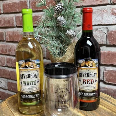 The Classics Gift Set, featuring a bottle of Riverboat white wine and a bottle of Riverboat red wine, is elegantly displayed on a wooden table. Between the bottles stands a clear tumbler adorned with the text "Bluff Top & Rocheport." A small pine branch with pinecones adds a touch of rustic charm behind the bottles, all set against a brick wall backdrop.