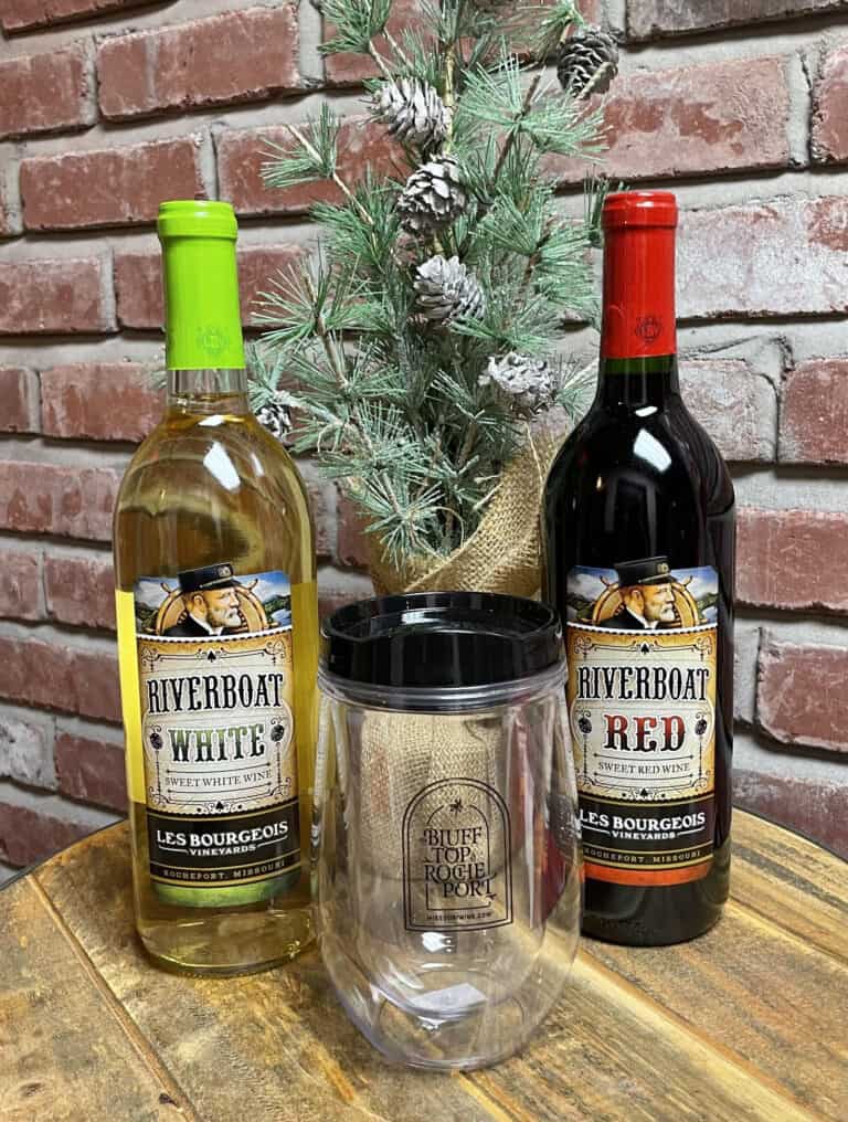 The Classics Gift Set, featuring a bottle of Riverboat white wine and a bottle of Riverboat red wine, is elegantly displayed on a wooden table. Between the bottles stands a clear tumbler adorned with the text 