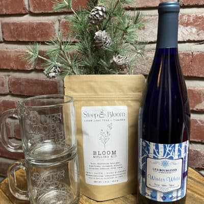 The Seasons Greetings Gift Set features a bottle of Winter White wine and a Steep & Bloom mulling kit. It includes two clear mugs adorned with text, artfully arranged beside the items. A sprig of pine with cones adds a festive touch to the display, set against a rustic brick wall backdrop.