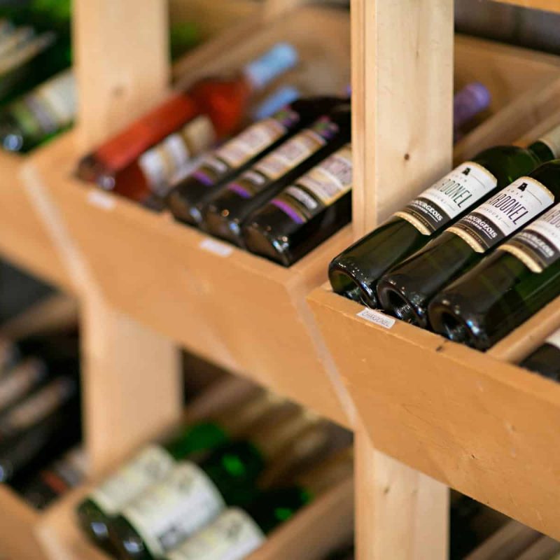 Wooden wine rack with several bottles of wine, including both red and white varieties, neatly arranged in angled compartments. The bottles have colorful labels and are stored in a cozy, rustic setting.