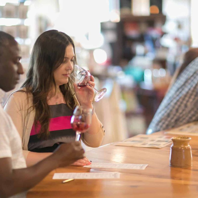 A man and woman sit at a bar, each holding a glass of red wine. The woman sips her wine while the man looks ahead. Other patrons can be seen in the background, with a tip jar and cash on the bar counter in the foreground.