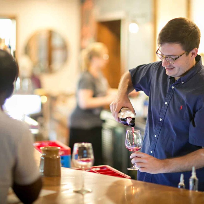 A man in a button-up shirt pours red wine into a glass at a bar. Two patrons, one in a white shirt, are seated across from him. In the background, a woman and bar decor are visible, creating a warm, welcoming atmosphere.