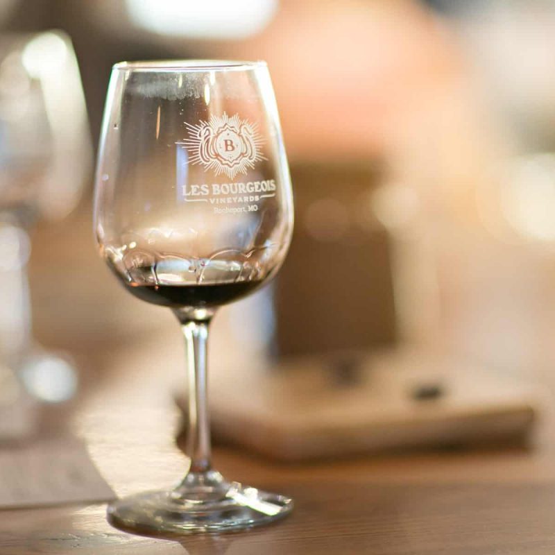 A wine glass partially filled with red wine sits on a wooden table. The glass has "LES BOURGEOIS VINEYARDS Rocheport, MO" written on it. The background is blurred with warm lighting, creating a cozy atmosphere.