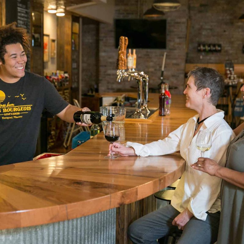 A bartender pours wine for two women seated at a wooden counter in a cozy bar setting. The bartender wears a t-shirt with a vineyard logo, while the women, holding wine glasses, appear engaged and content. The bar interior features brick walls and various bottles.