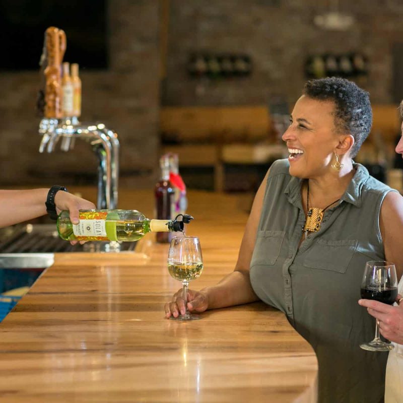 Two smiling women stand at a bar counter, one holding a glass of white wine while the bartender pours more wine into it. The other woman holds a glass of red wine. The background includes a beer tap and shelves with bottles.