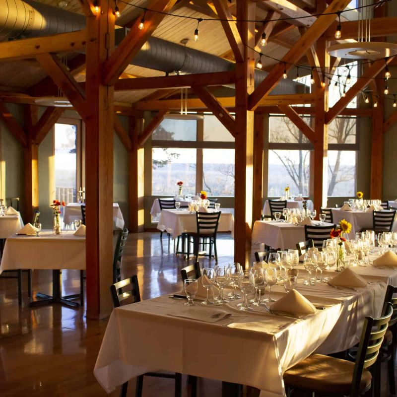 A warmly lit restaurant with large windows and wooden beams, showcasing neatly arranged tables with white tablecloths, folded napkins, and flower centerpieces. Empty chairs surround the tables, and string lights hang from the ceiling, adding a cozy ambiance.