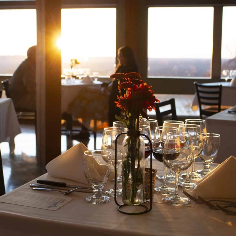 A restaurant table set with glassware, white napkins, and a vase with red flowers is bathed in warm sunset light. Large windows offer a scenic view. In the background, a couple is seated at another table, enjoying the serene atmosphere.