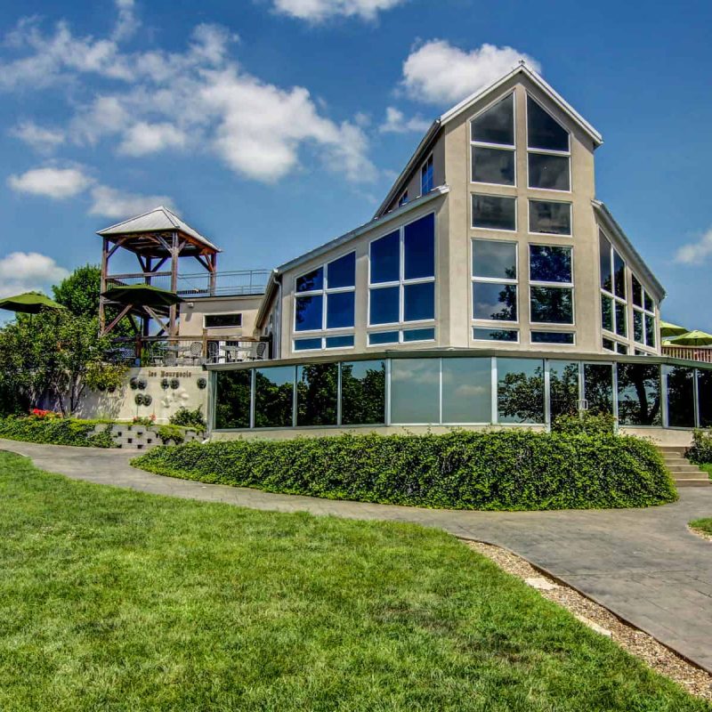 A modern, multi-story building with large glass windows and solar panels, surrounded by a lush green lawn. A stone path leads to the entrance. The structure includes a tall observation tower with a roof, under a partly cloudy blue sky.