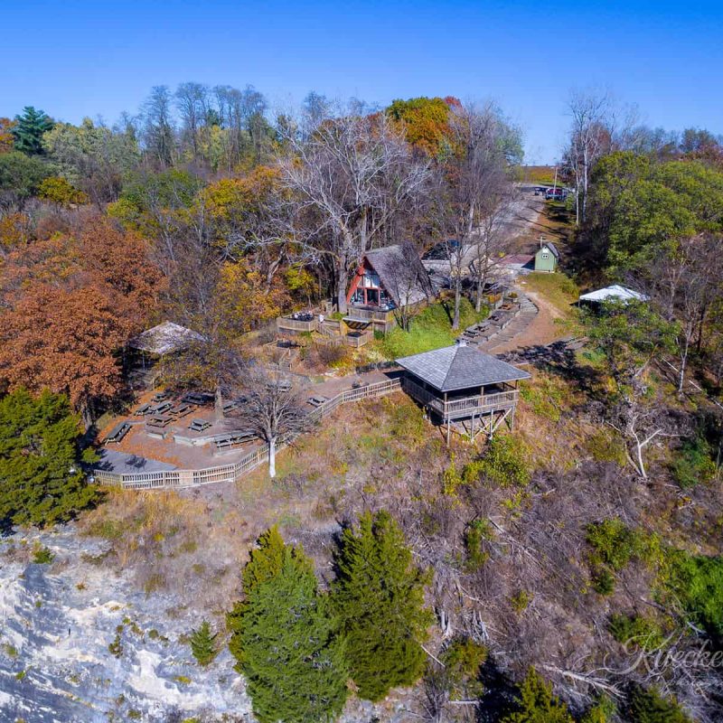 Aerial view of a tree-covered bluff with a wooden shelter and picnic tables spread across the area. The landscape features a mix of autumn and green foliage, with a clear blue sky overhead. There are pathways connecting different sections of the site.