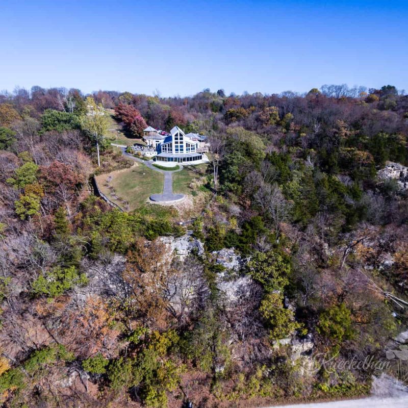 Aerial view of a large house with a modern design situated on a hill surrounded by dense forest. The house features large windows and appears to overlook a small clearing. Trees with autumn foliage fill the landscape, and a clear blue sky is above.