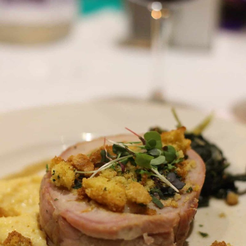 A plate of gourmet food featuring a rolled meat dish topped with breadcrumbs and garnished with microgreens, served with a side of creamy polenta and sautéed greens. A glass of red wine is in the background.