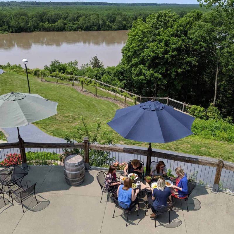 A group of people sitting around a table with food and drinks under blue umbrellas on an outdoor patio. The patio overlooks a river and lush green landscape with trees. Several empty tables and chairs are also visible on the patio.