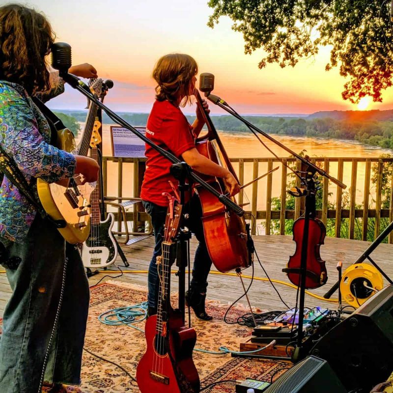 Three musicians perform on a wooden deck at sunset, overlooking a scenic river. One plays a guitar, another a cello, and the third a banjo. Various string instruments and microphones are set up around them. The vibrant sunset creates a warm and picturesque backdrop.