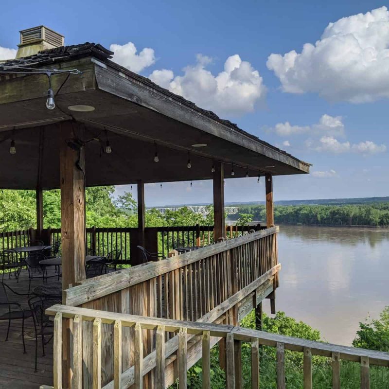 A wooden deck with a roof overlooks a river on a sunny day. The deck has a few empty tables and chairs. The surrounding area is lush with green trees, and the sky is partly cloudy with white fluffy clouds.