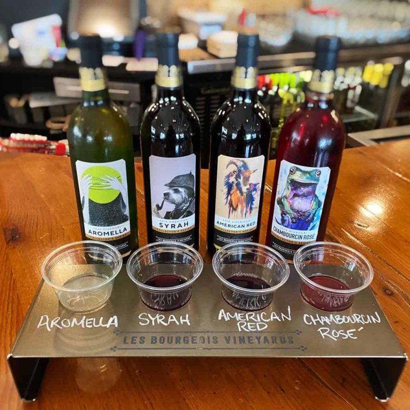 Four bottles of wine with colorful labels and tasting cups are arranged on a wooden table. The labels read Aromella, Syrah, American Red, and Chambourcin Rosé, from Les Bourgeois Vineyards. The glasses hold small samples of each wine in front of the corresponding bottle.