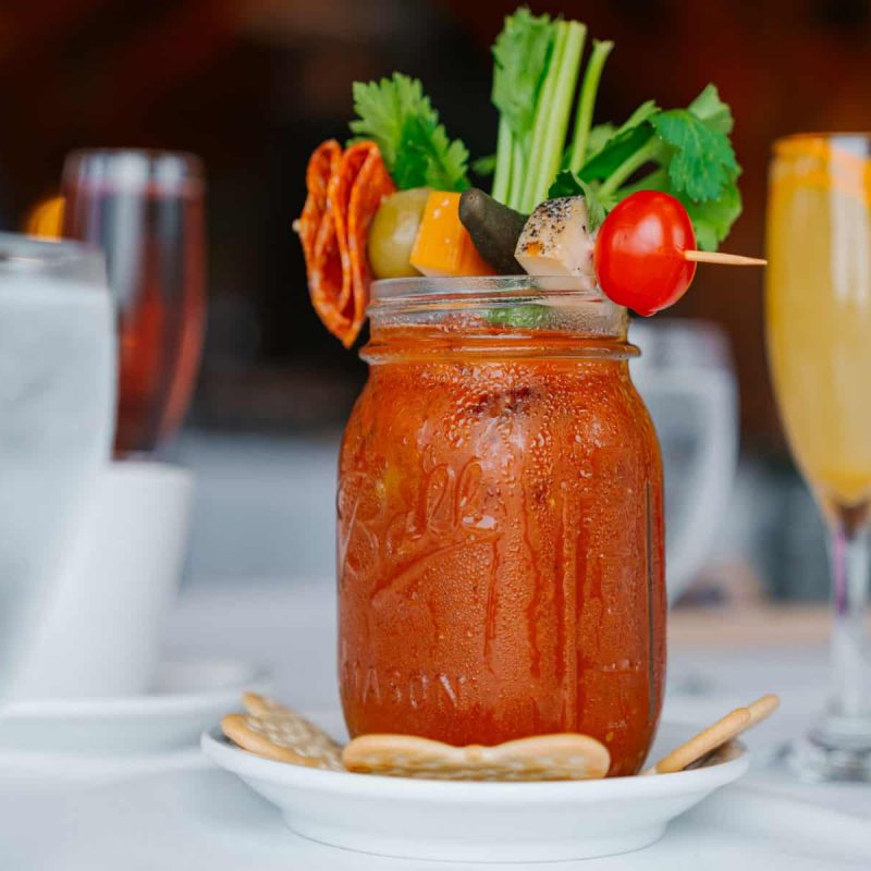 A refreshing Bloody Mary cocktail garnished with celery, a cherry tomato, olives, and a slice of meat in a mason jar sits on a white plate with crackers. Surrounding the drink are glasses of water and another beverage garnished with an orange slice.