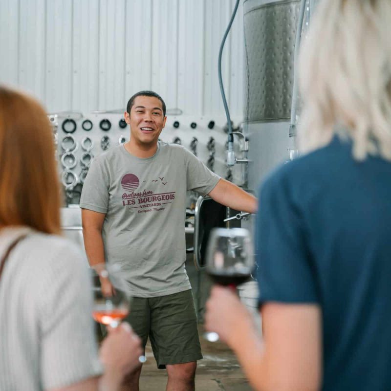 A man in a casual outfit, standing near large metal tanks, smiles and talks to two people. The people listening, one with red hair and the other with blonde hair, hold wine glasses in their hands inside a winery.