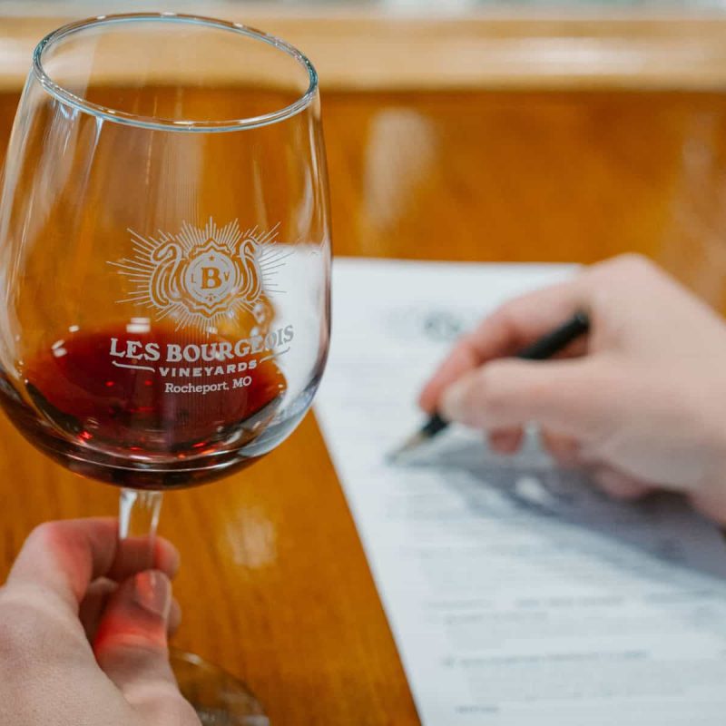 A person holds a glass of red wine with "Les Bourgeois Vineyards Rocheport, MO" printed on it. Their other hand is holding a pen and writing on a sheet of paper placed on a wooden surface.