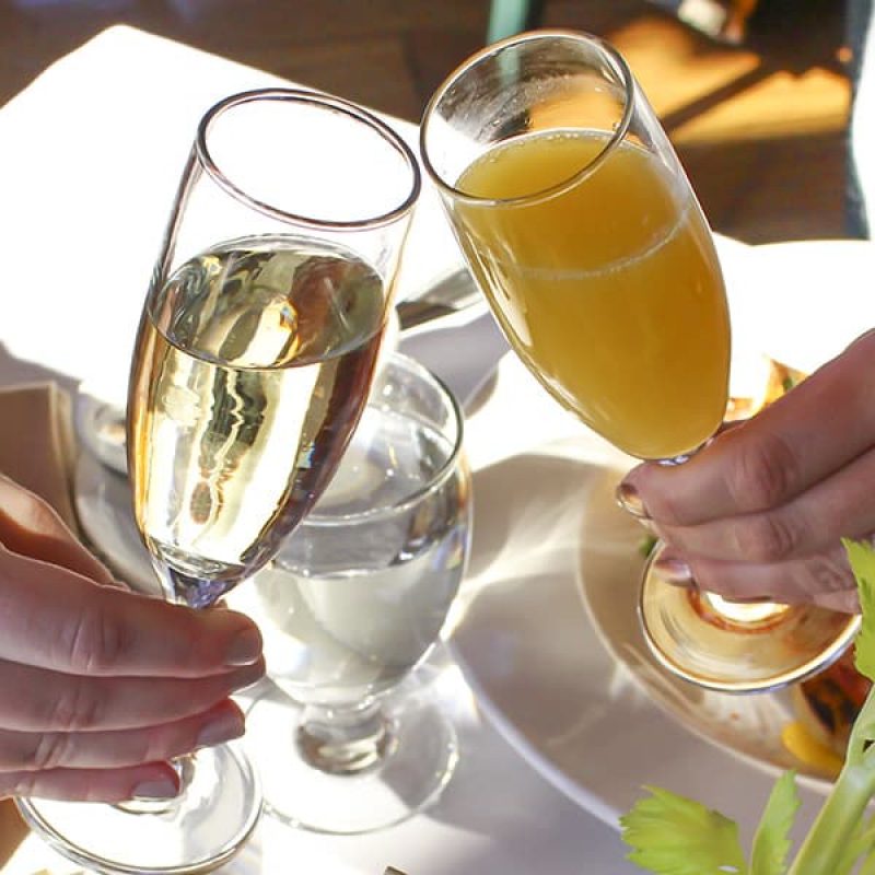 Two people clinking glasses, one with a bubbly champagne or prosecco and the other with orange mimosa, over a table set with a glass of water and other items in the background. The image is brightly lit, suggesting a daytime setting.