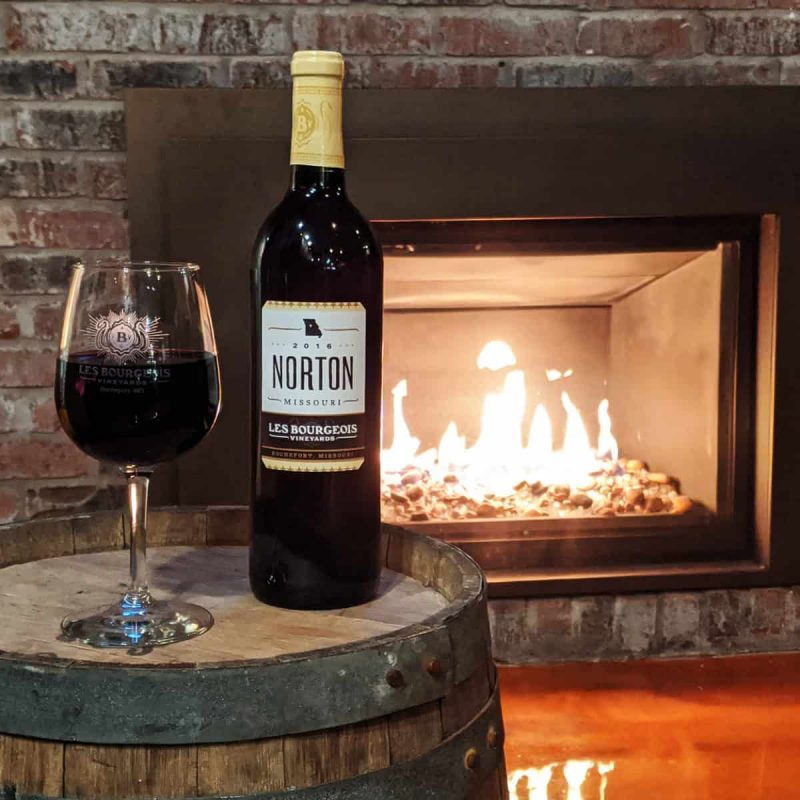 A bottle of Norton wine and a glass of red wine sit on a wooden barrel in front of a lit fireplace encased in a brick wall. The warm glow from the fire enhances the cozy atmosphere.