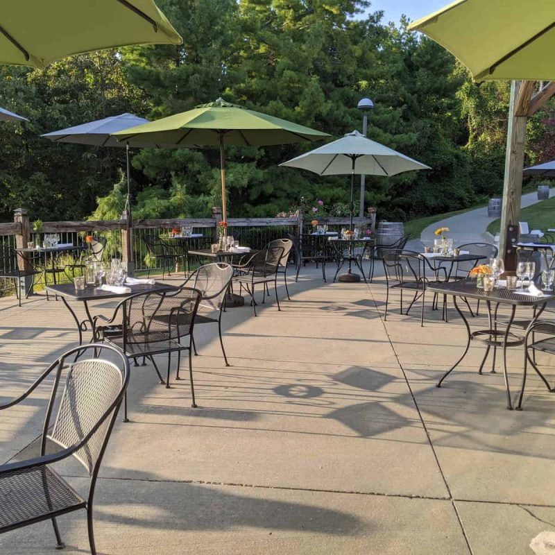 Outdoor patio dining area with numerous metal tables and chairs, partially shaded by large umbrellas. Each table is set with glasses and small flower vases. The patio is surrounded by greenery, contributing to a serene and inviting atmosphere.