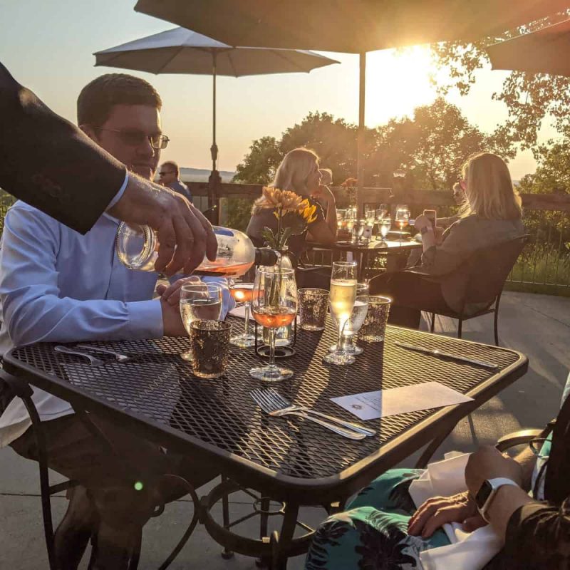 A man pours water into a glass at an outdoor restaurant during sunset. Other patrons are seated at tables, with umbrellas providing shade. The scene is warm and inviting, with sunlight filtering through trees and casting a golden glow.