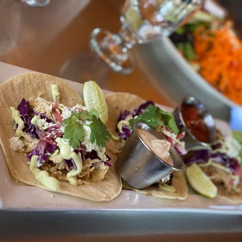 Three fish tacos topped with shredded purple cabbage, cilantro, and sliced radishes are arranged on a rectangular white plate. A small metal cup of sauce and lime wedges accompany the tacos. Additional dishes, including a salad, are visible in the background.