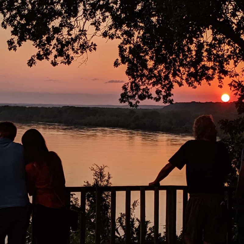 An orange sunset over the river. Four people in silhouette admire the view from the A-Frame deck